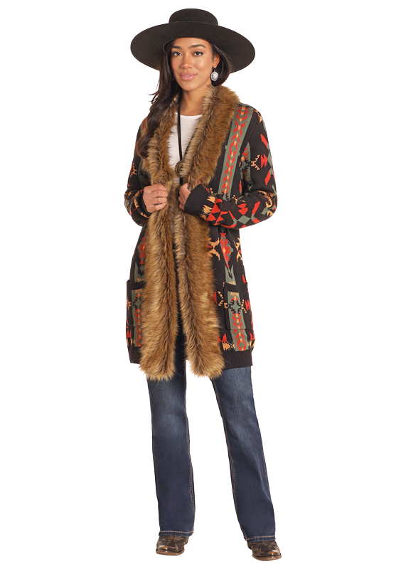 A woman wearing a multicolored cardigan with a geometric Aztec pattern and a plush fur trim on the hood and pockets. She is also wearing a wide-brimmed hat and boots, and the setting appears to be a studio shot.