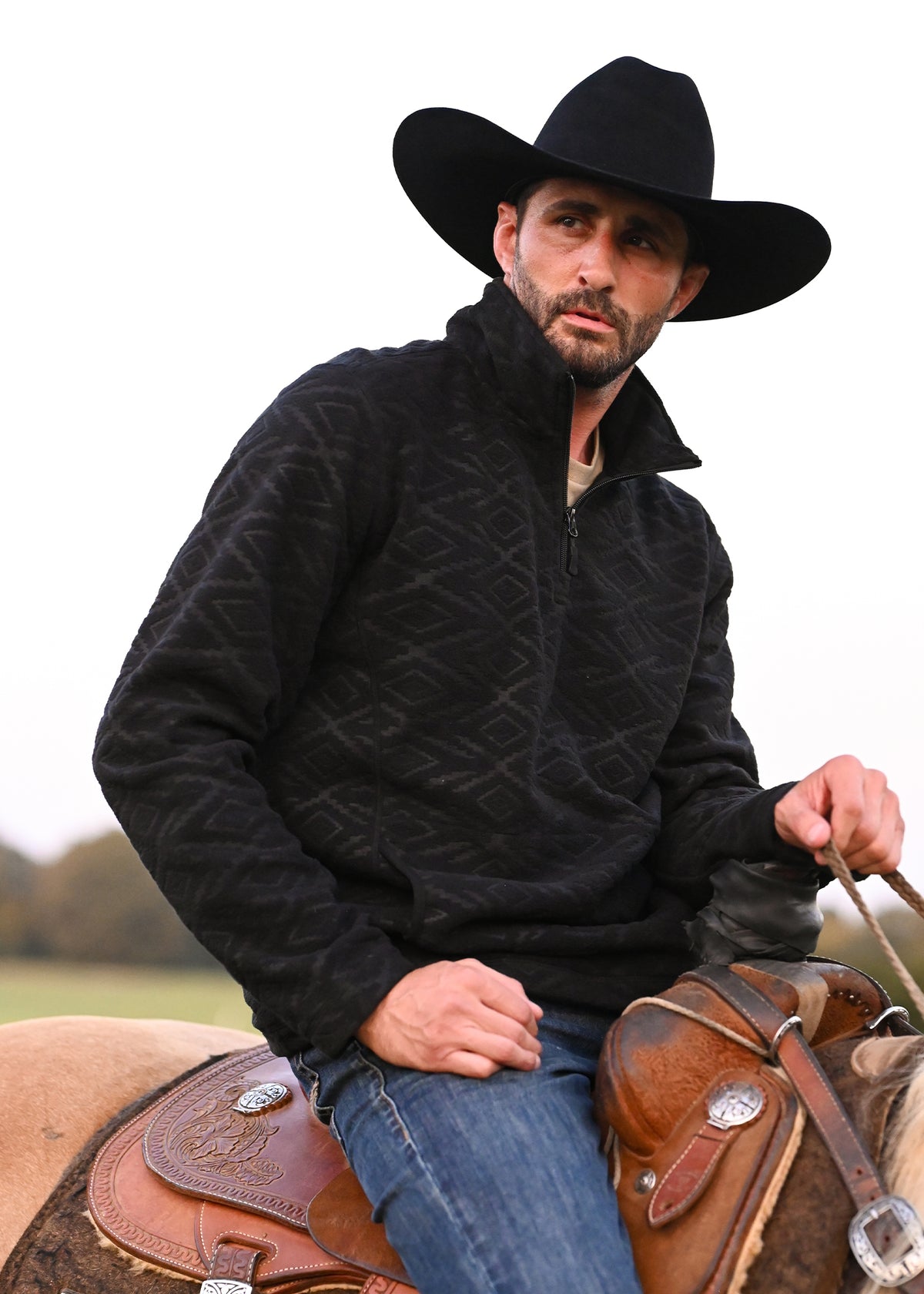 A man wearing a black cowboy hat and a black textured quarter-zip fleece jacket sits on a horse saddle outdoors.