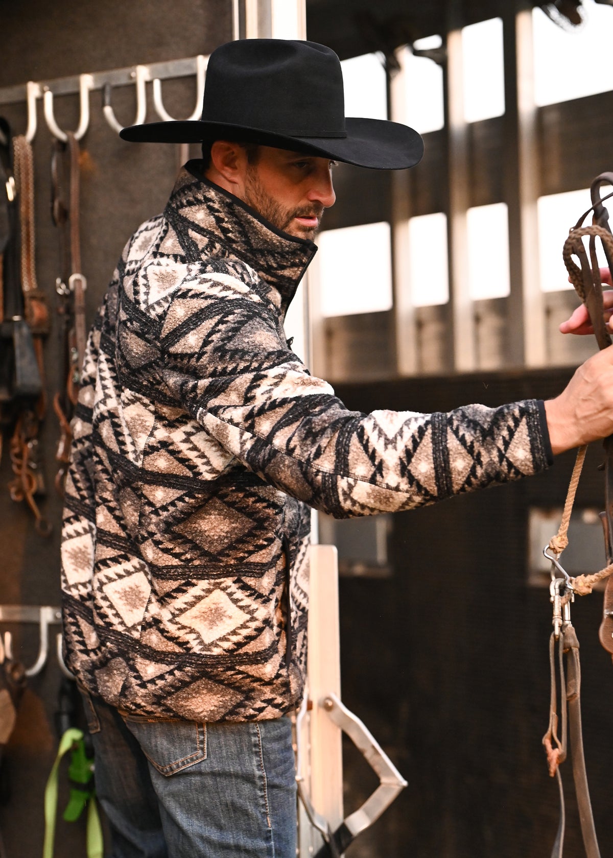 A man wearing a black cowboy hat and a beige, black, and brown geometric patterned fleece jacket stands in a rustic barn setting, holding horse tack.