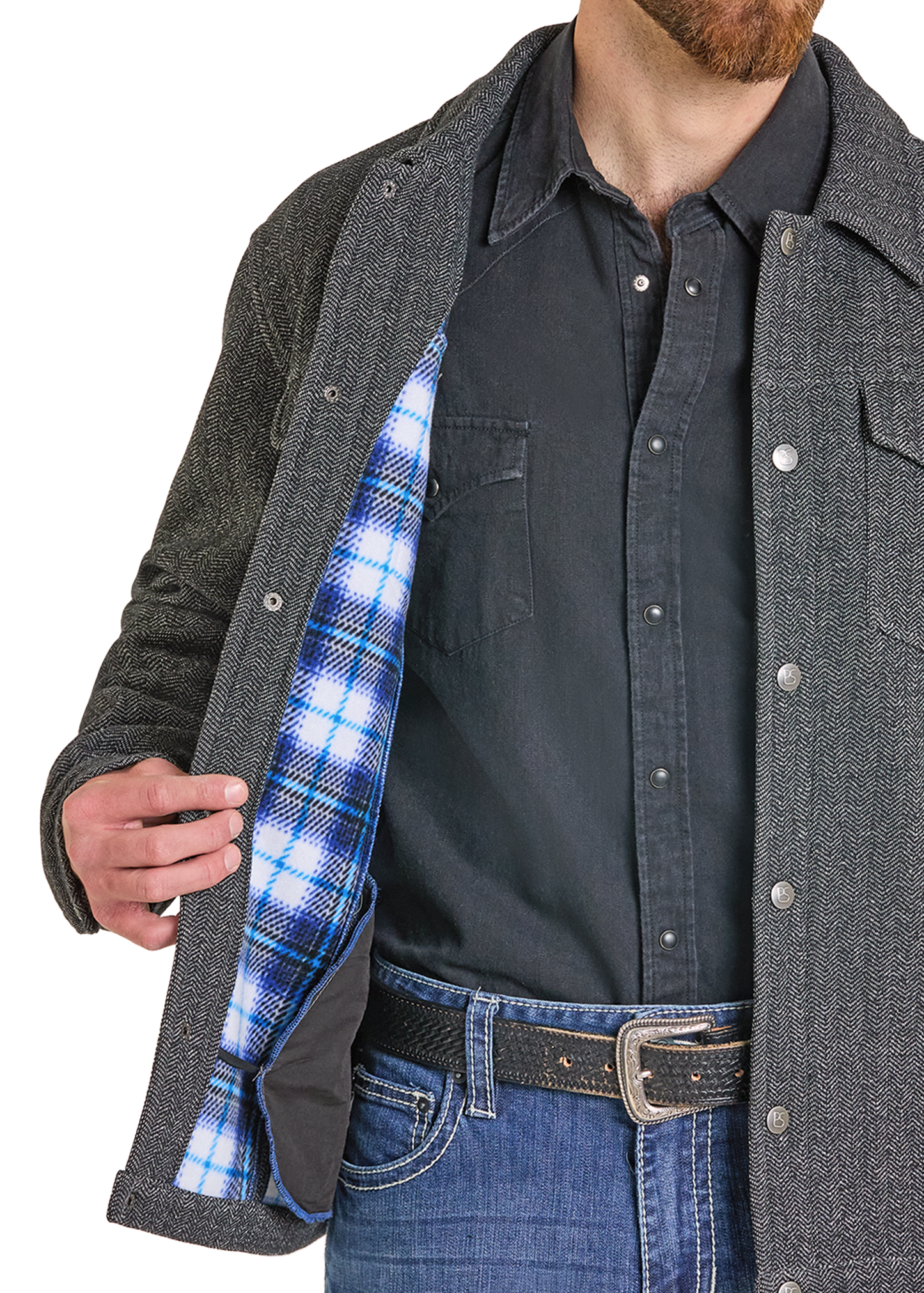 A close-up of a man wearing a dark gray textured overshirt over a black button-up shirt and blue jeans, opening the overshirt to reveal a blue and white plaid lining.