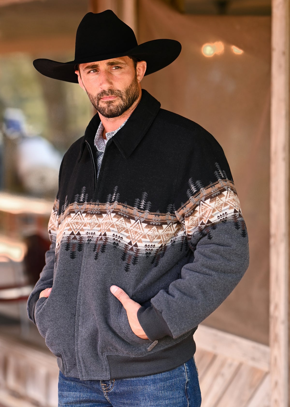 A man wearing a black cowboy hat and a dark gray fleece zip-up jacket with a brown and beige geometric pattern across the chest, standing in front of a rustic wooden building.
