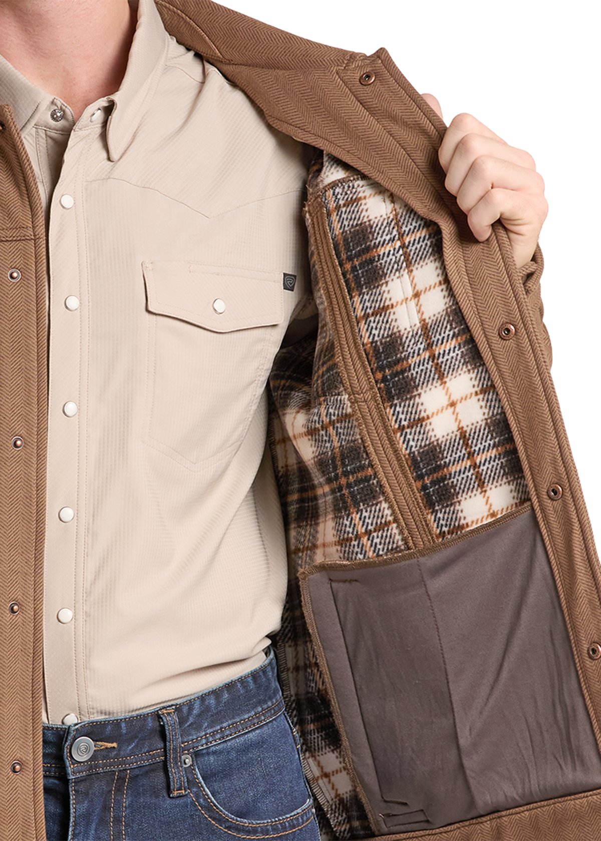 A close-up of a person wearing a tan overshirt over a beige button-up shirt and blue jeans, opening the overshirt to show a brown zippered pocket and a plaid-lined interior.