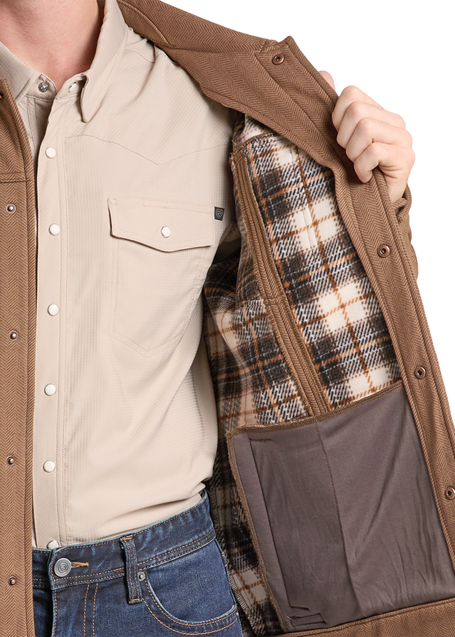 A close-up of a person wearing a tan overshirt over a beige button-up shirt and blue jeans, opening the overshirt to show a brown zippered pocket and a plaid-lined interior.