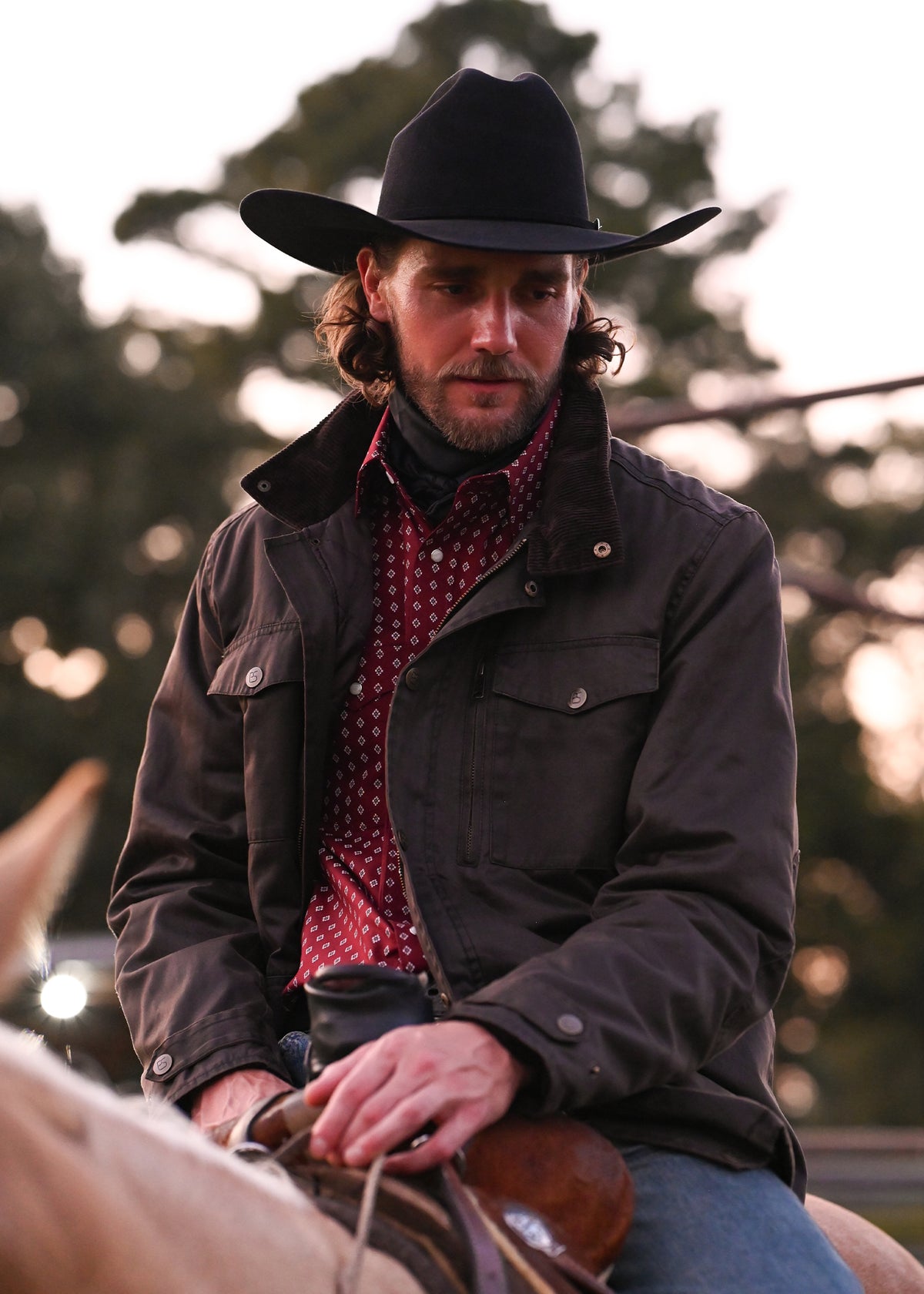 A man wearing a black cowboy hat, a dark brown jacket, and a red patterned shirt while riding a horse outdoors at sunset.