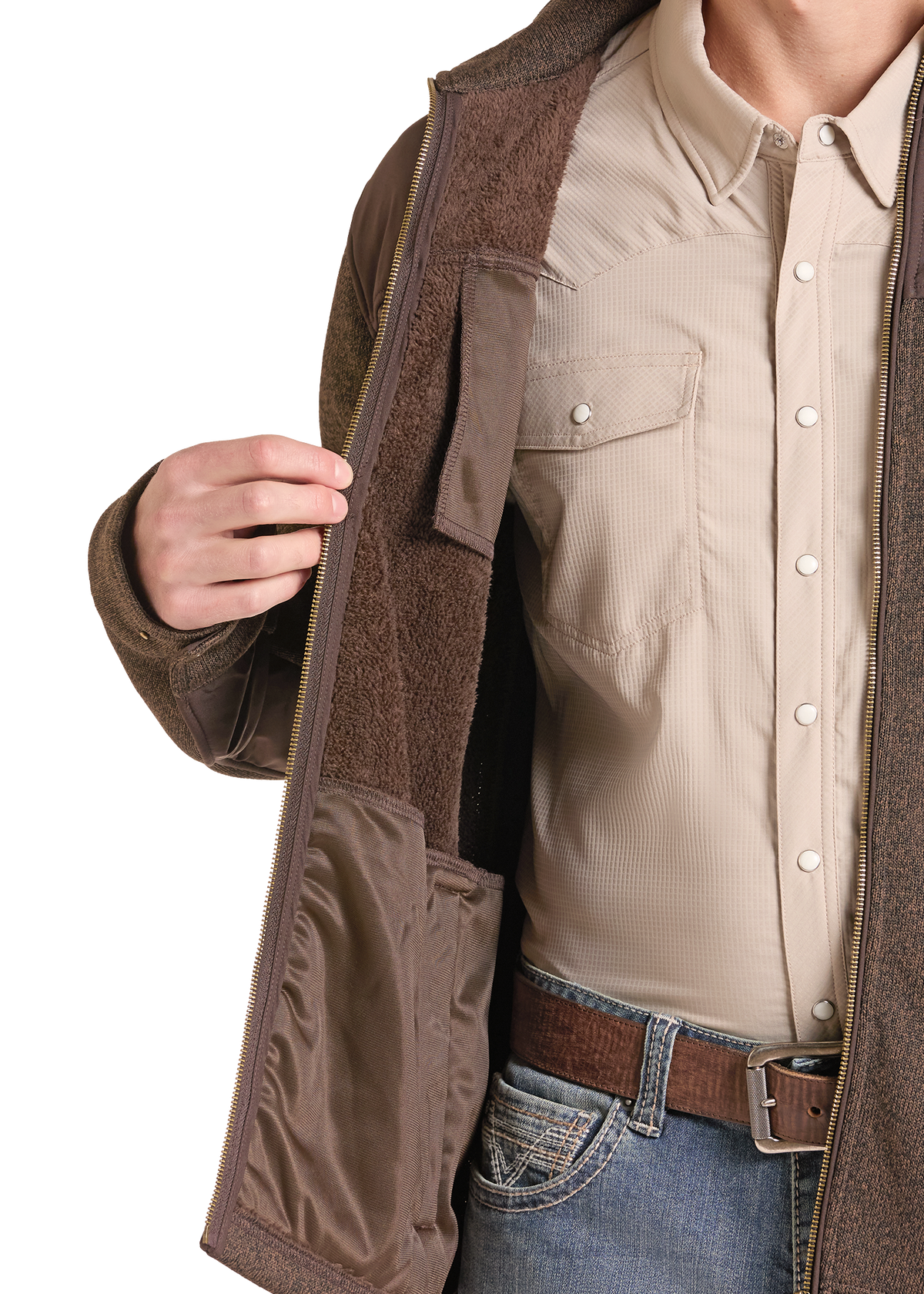 A close-up of a person wearing a brown zip-front jacket with a brown sherpa lining over a beige button-up shirt and blue jeans.