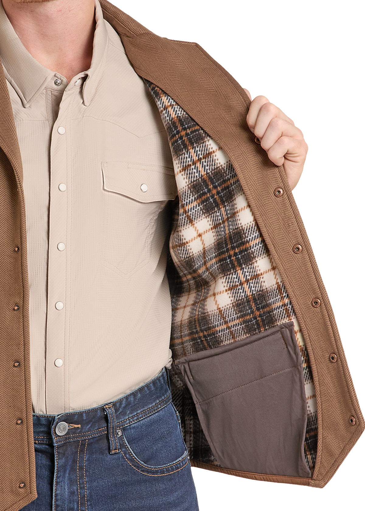 A close-up of a person wearing a tan overshirt over a beige button-up shirt and blue jeans, opening the overshirt to show a brown plaid lining and a brown interior pocket.