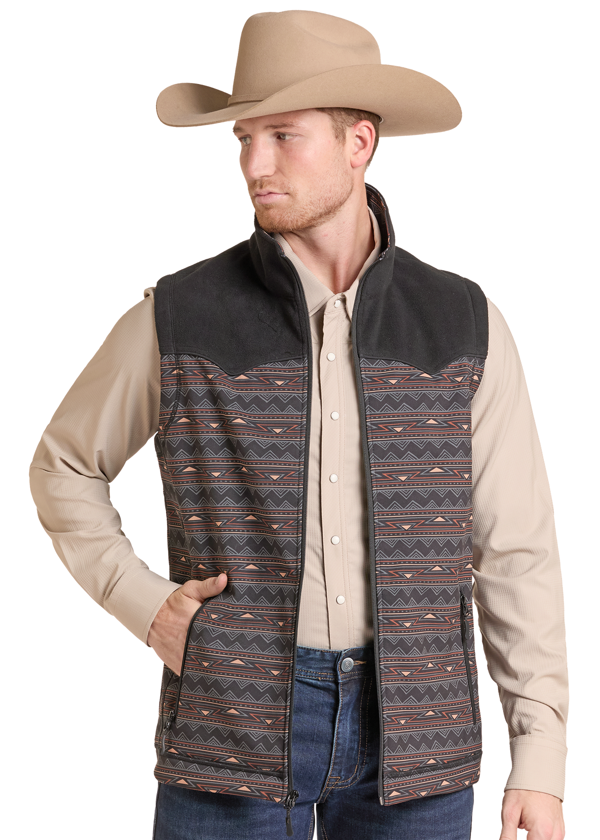 A man wearing a tan cowboy hat, beige long-sleeve shirt, and a western-style vest with a dark suede-like yoke and a geometric patterned body, paired with blue jeans.