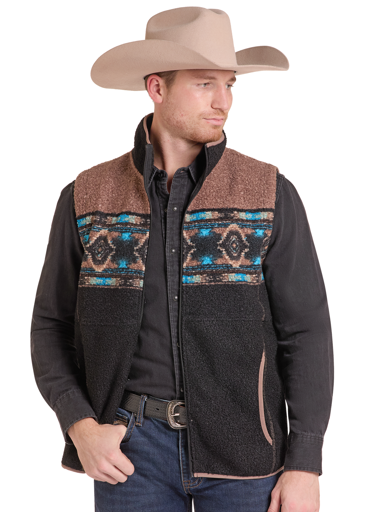 A man wearing a brown and black western vest with a colorful geometric pattern across the chest, a black long-sleeve shirt, blue jeans, and a beige cowboy hat.