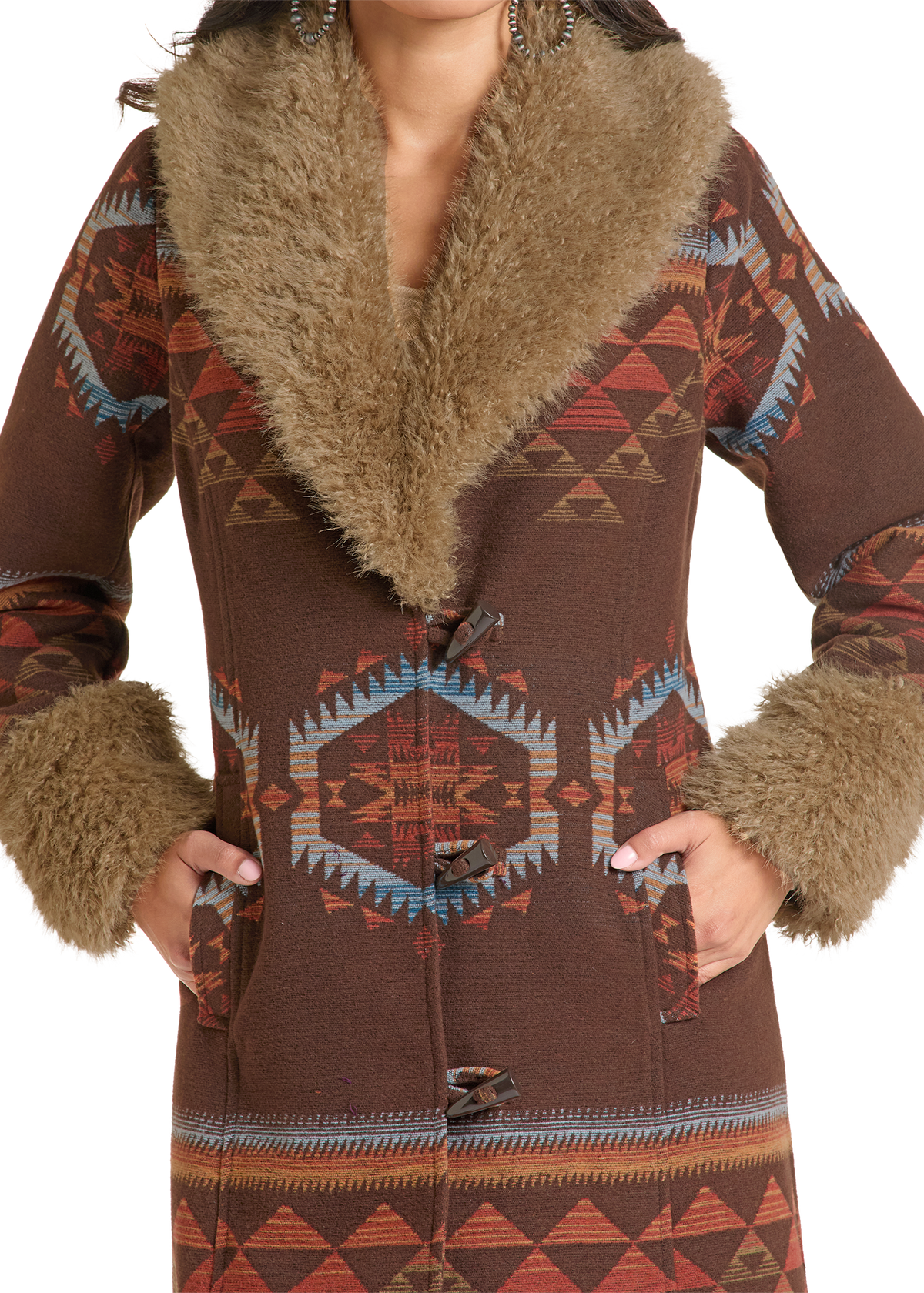 A close-up of a woman wearing a brown patterned coat with a large tan faux-fur collar and matching faux-fur trim at the cuffs and pockets. The coat features a Southwestern geometric pattern in brown, rust, blue, and cream.