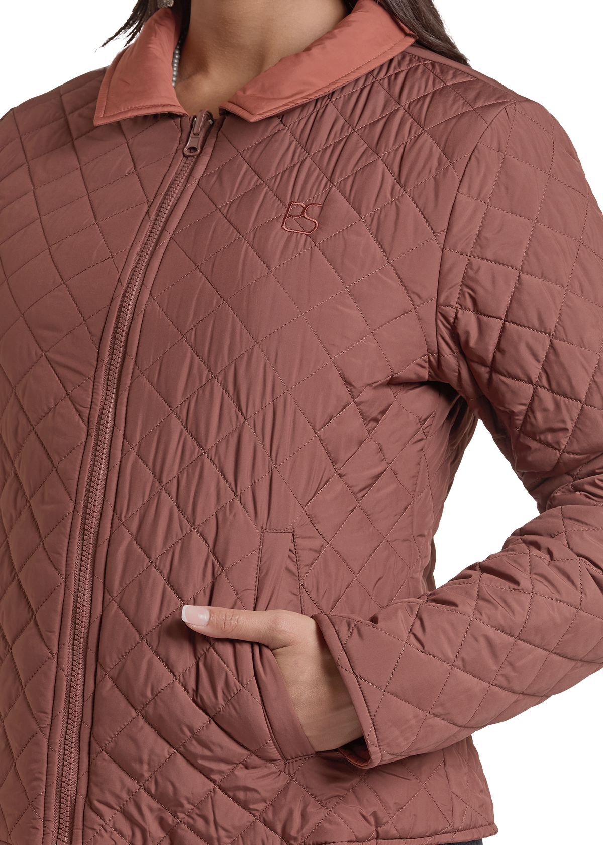 A close-up of a person wearing a quilted zip-front jacket in a dusty rose color, with a small embroidered logo on the chest and a hand tucked into a side pocket.