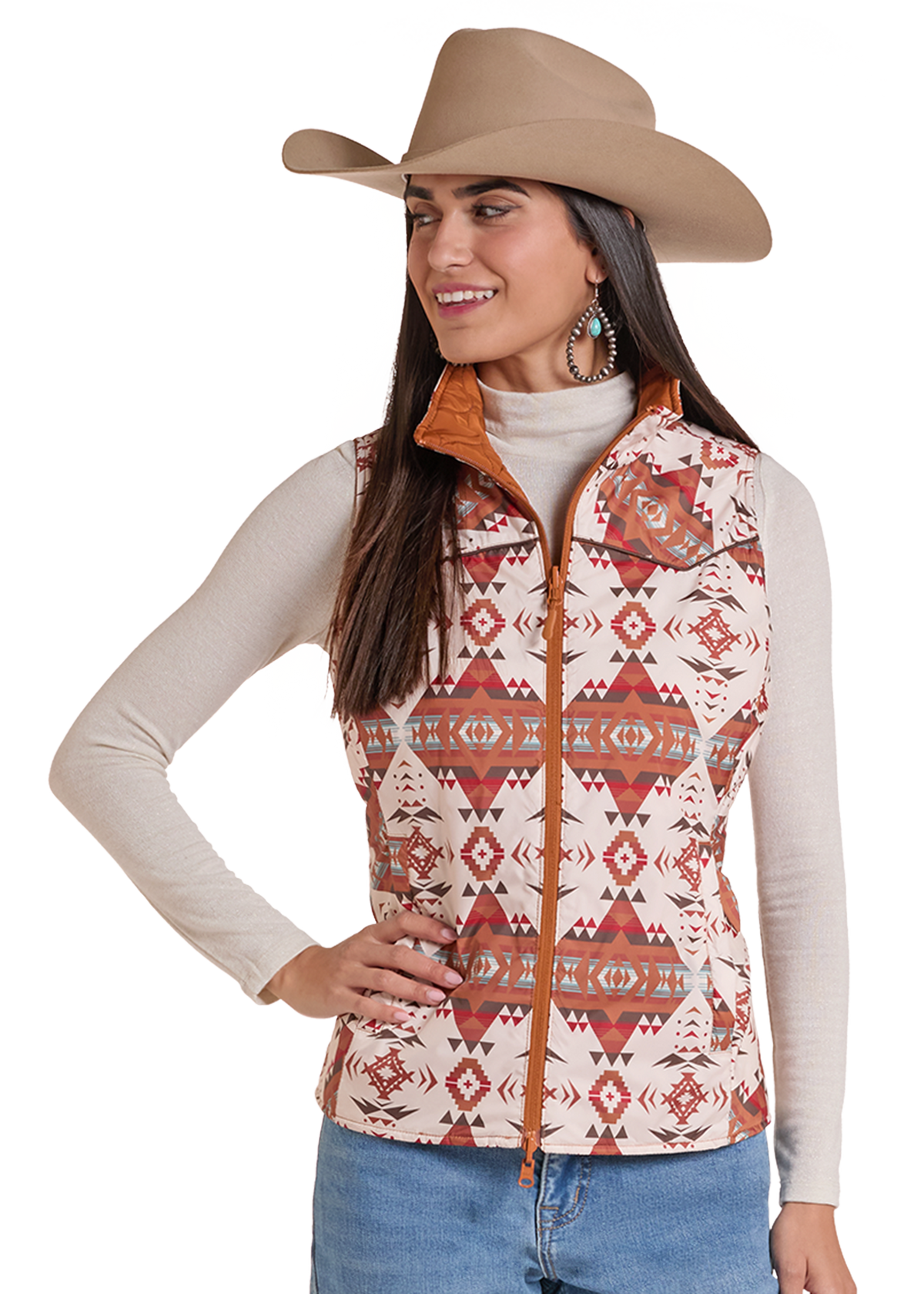 A woman wearing a beige cowboy hat, a cream long-sleeve shirt, and a sleeveless western vest with a red, orange, and beige Southwestern geometric pattern, paired with blue jeans.