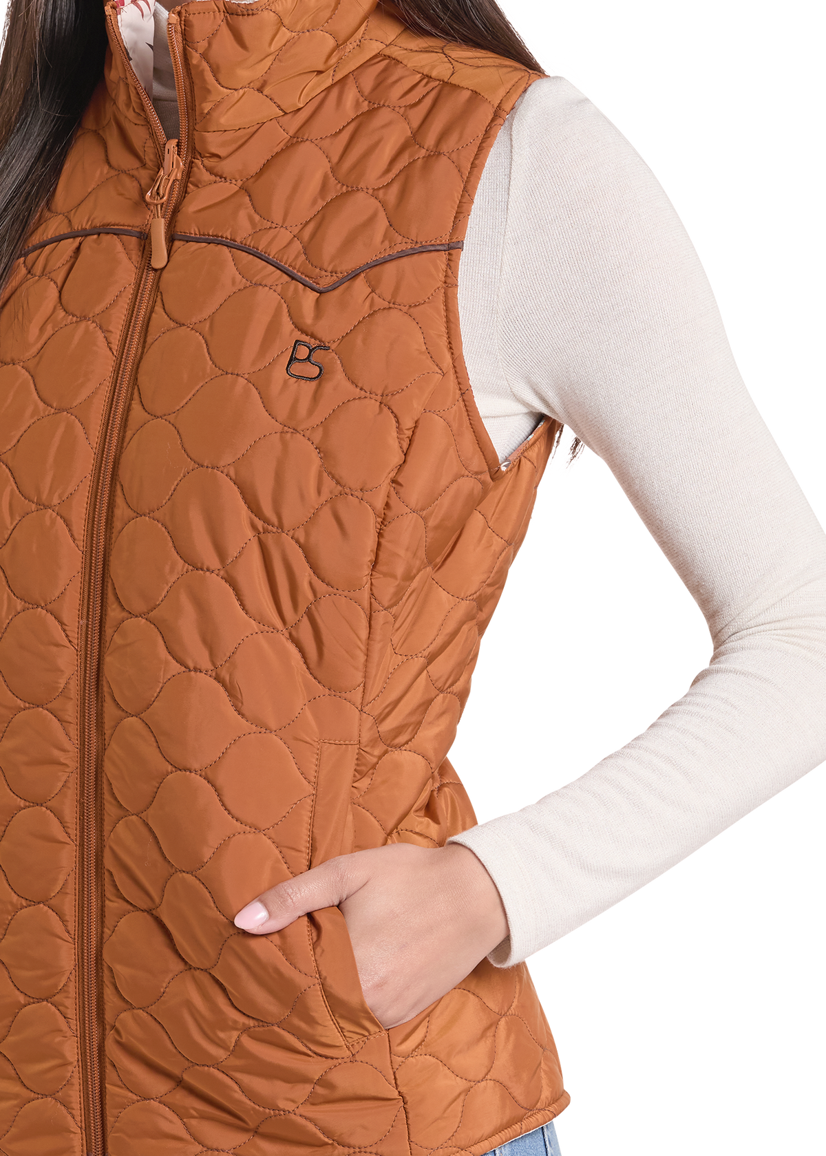 A close-up of a woman wearing a rust-brown quilted sleeveless puffer vest over a cream long-sleeve shirt. The vest features a full zip, a stand collar, and a small embroidered logo on the left chest.