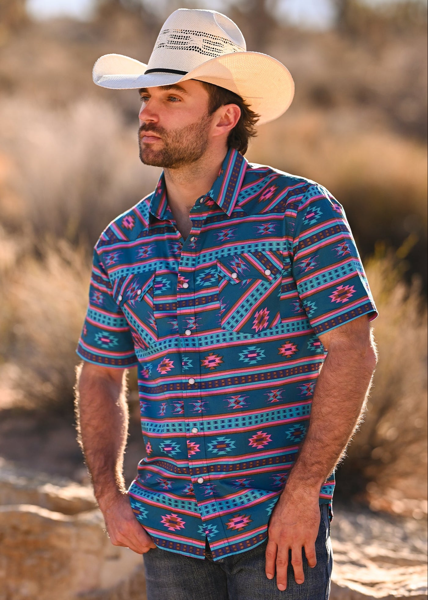 Man wearing a patterned shirt and cowboy hat in a desert setting