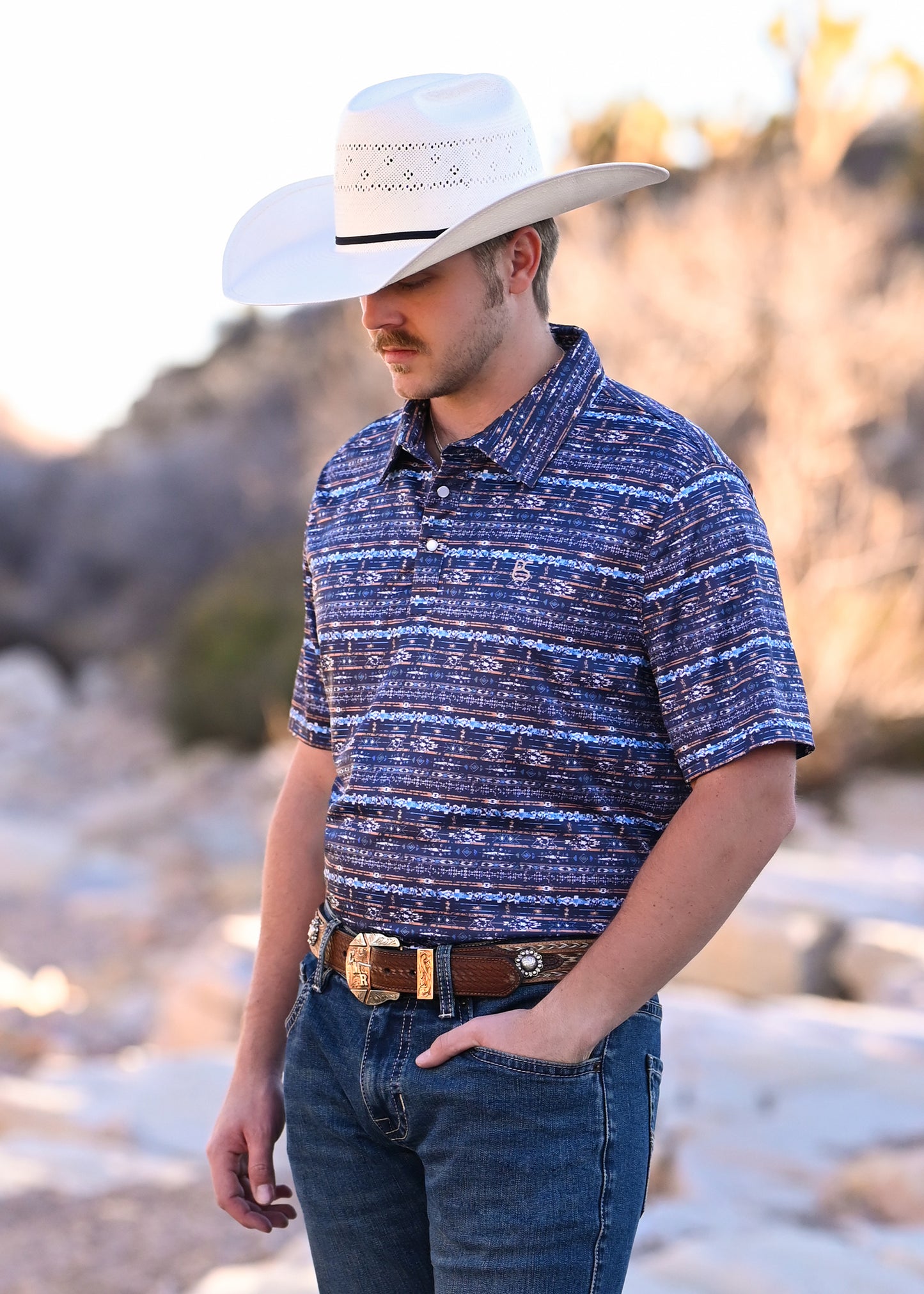 Man wearing a blue patterned shirt, white cowboy hat, and jeans standing outdoors.
