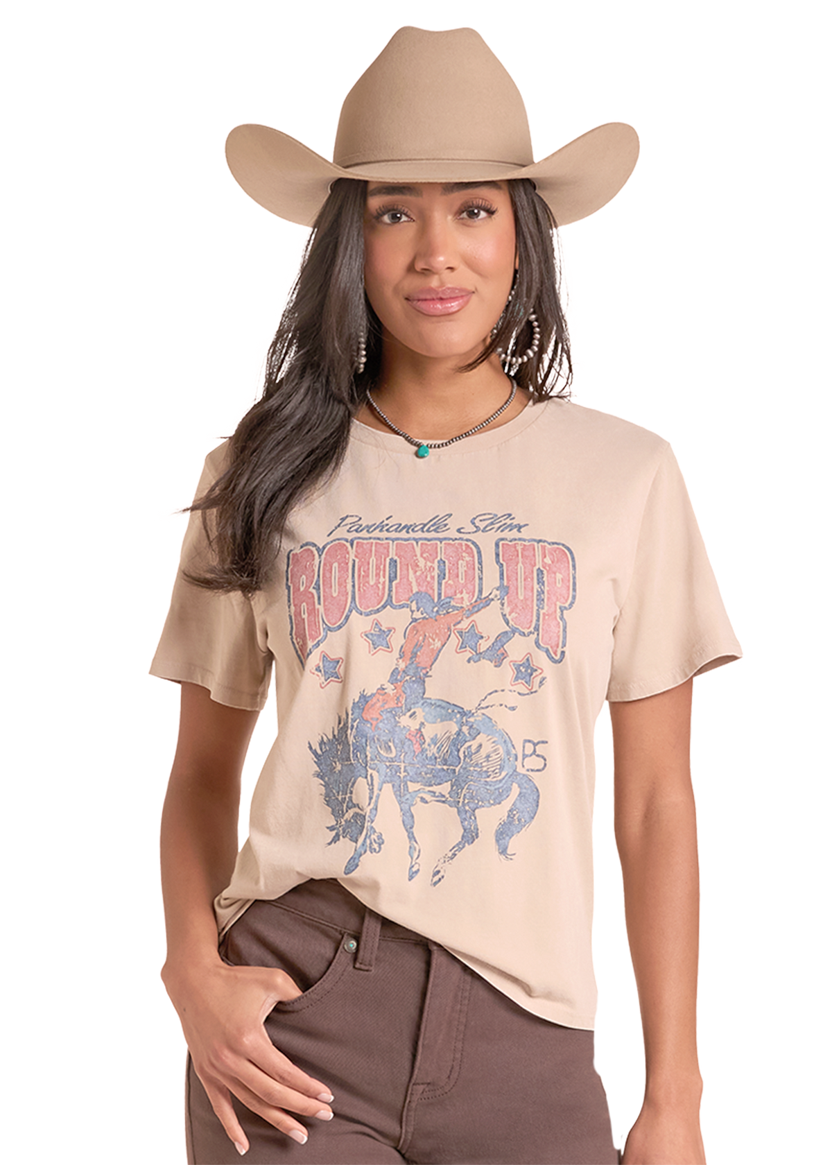A woman modeling a beige graphic t-shirt with a western theme, featuring a cowboy hat, brown pants, and a turquoise necklace.