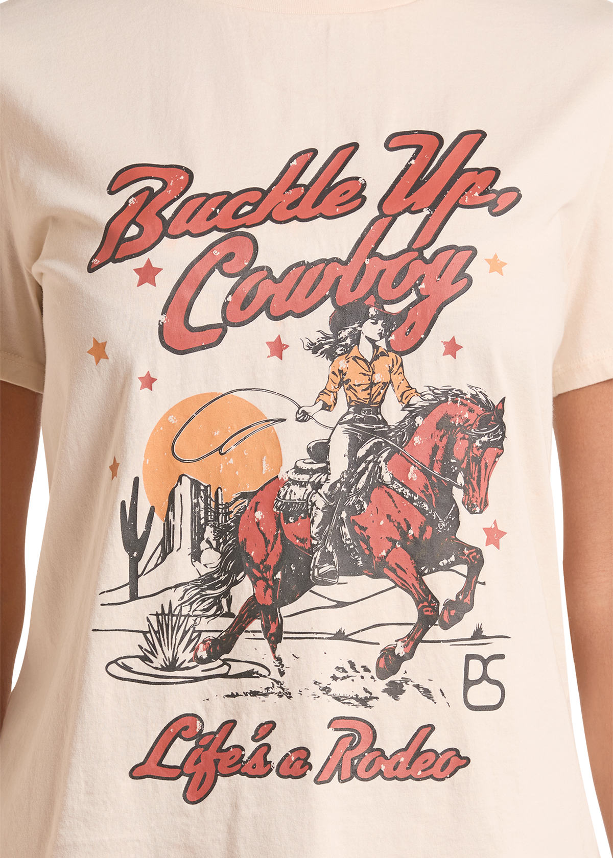 A close-up of a cream-colored t-shirt featuring a western-themed graphic print with the text 'Buckle Up, Cowboy' and 'Life's a Rodeo'. The graphic depicts a cowboy riding a red horse under a large orange sun, with desert cacti and stars.