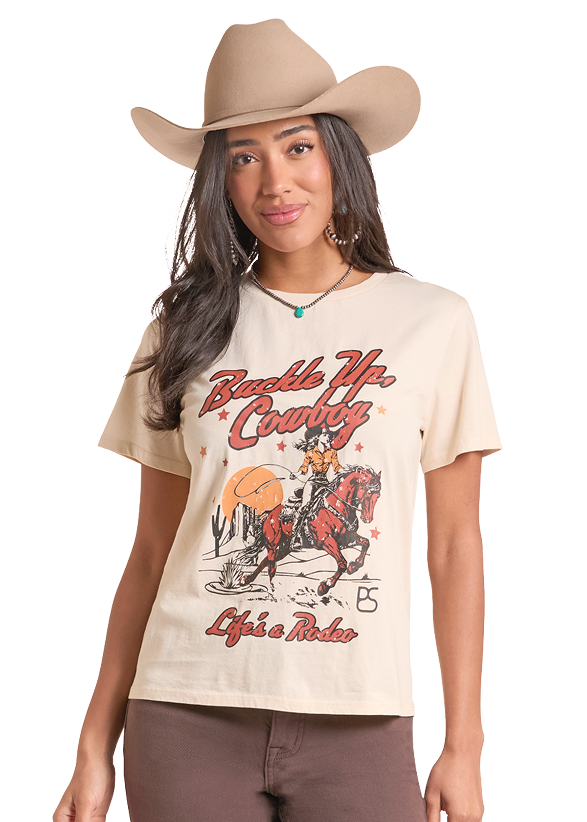 A woman wearing a beige graphic t-shirt with a western 'Buckle Up, Cowboy' design, paired with a matching beige cowboy hat and brown pants.