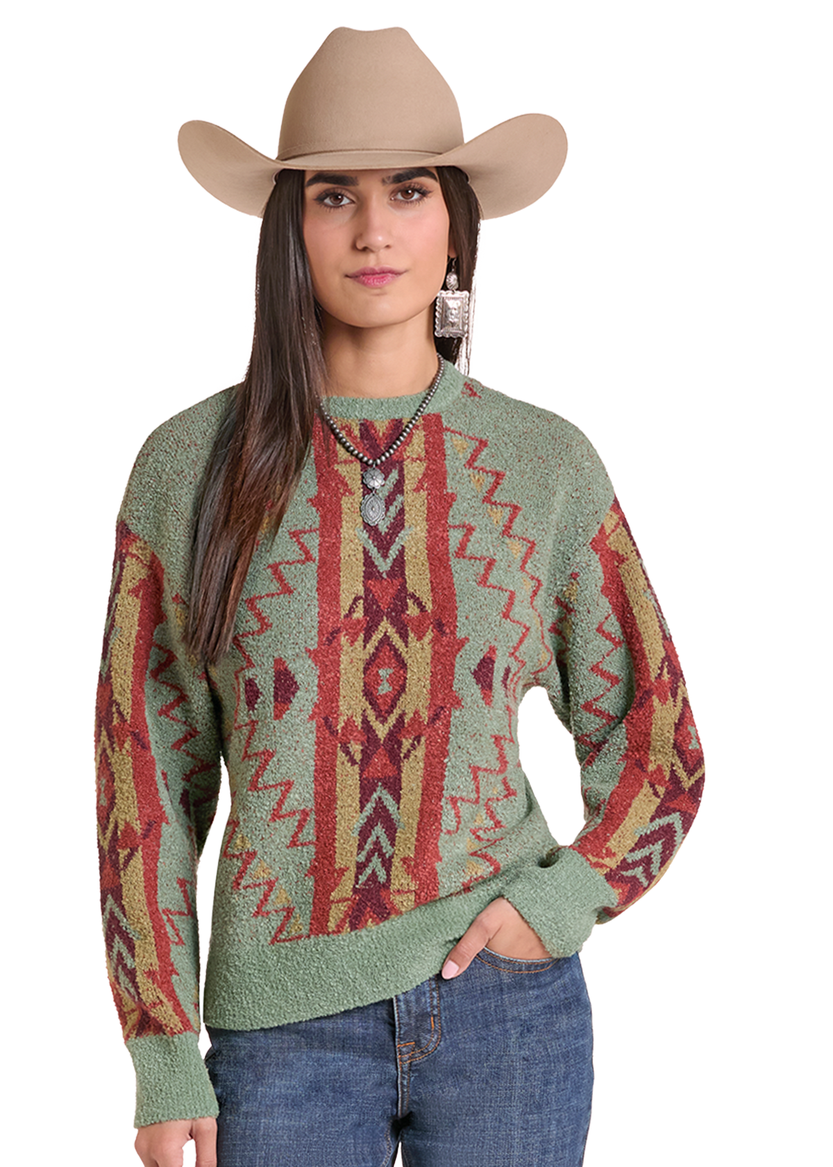A woman modeling a fuzzy knit long-sleeve sweater with a southwestern geometric pattern in sage green, rust red, and tan, paired with a beige cowboy hat and blue jeans.