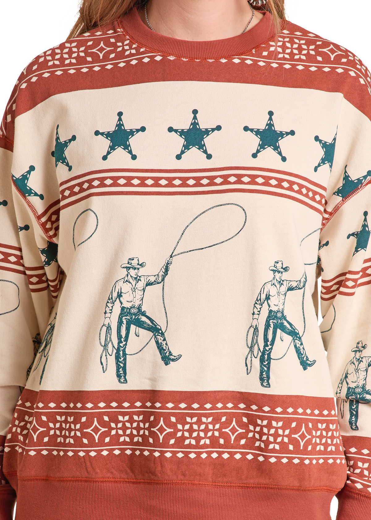A close-up of a crewneck sweatshirt featuring a western theme. The sweatshirt has a cream base with rust red borders and teal blue cowboy illustrations and star motifs.