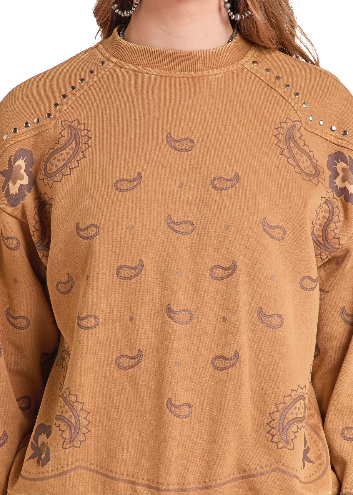 A close-up of a person wearing a tan crewneck sweatshirt with a brown paisley pattern and silver stud embellishments along the shoulders.