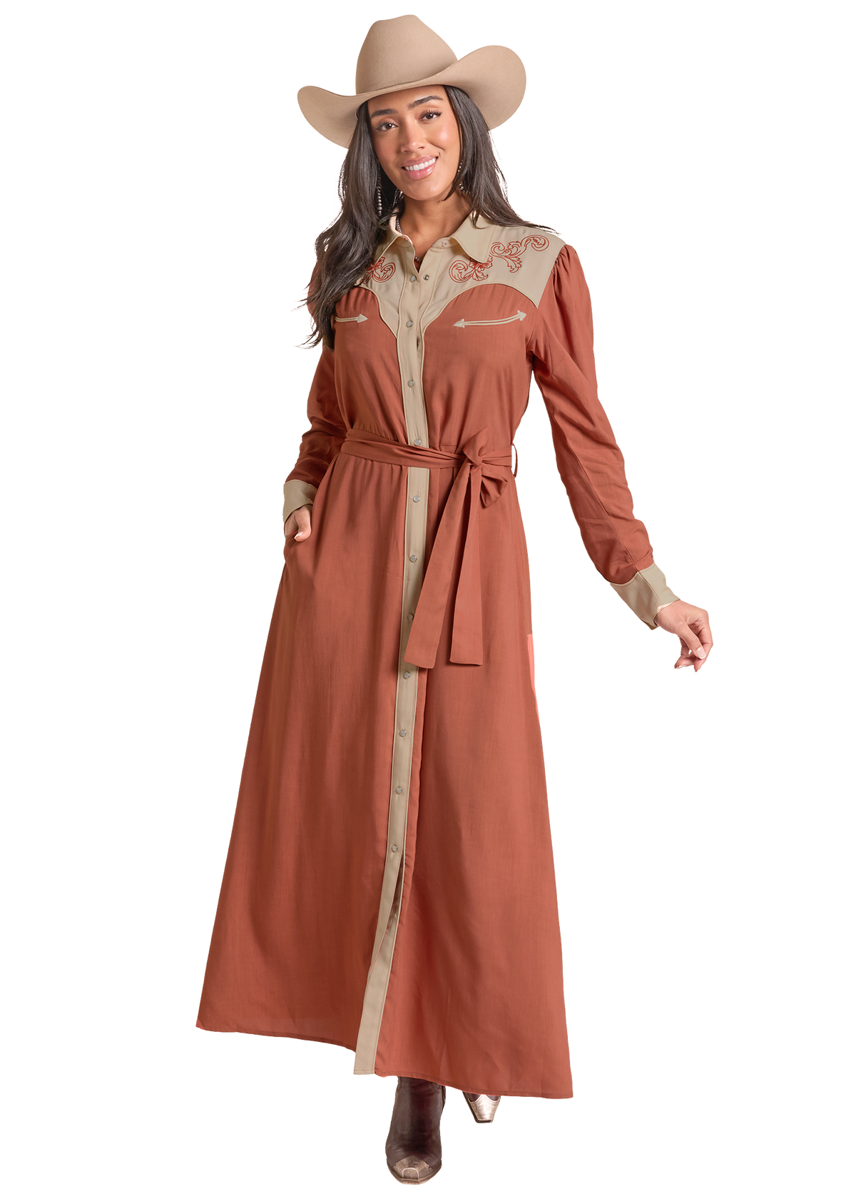 A woman modeling a long rust-brown western-style maxi shirt dress with beige contrast panels, embroidery on the chest, and a matching waist tie, paired with a tan cowboy hat against a plain white background.