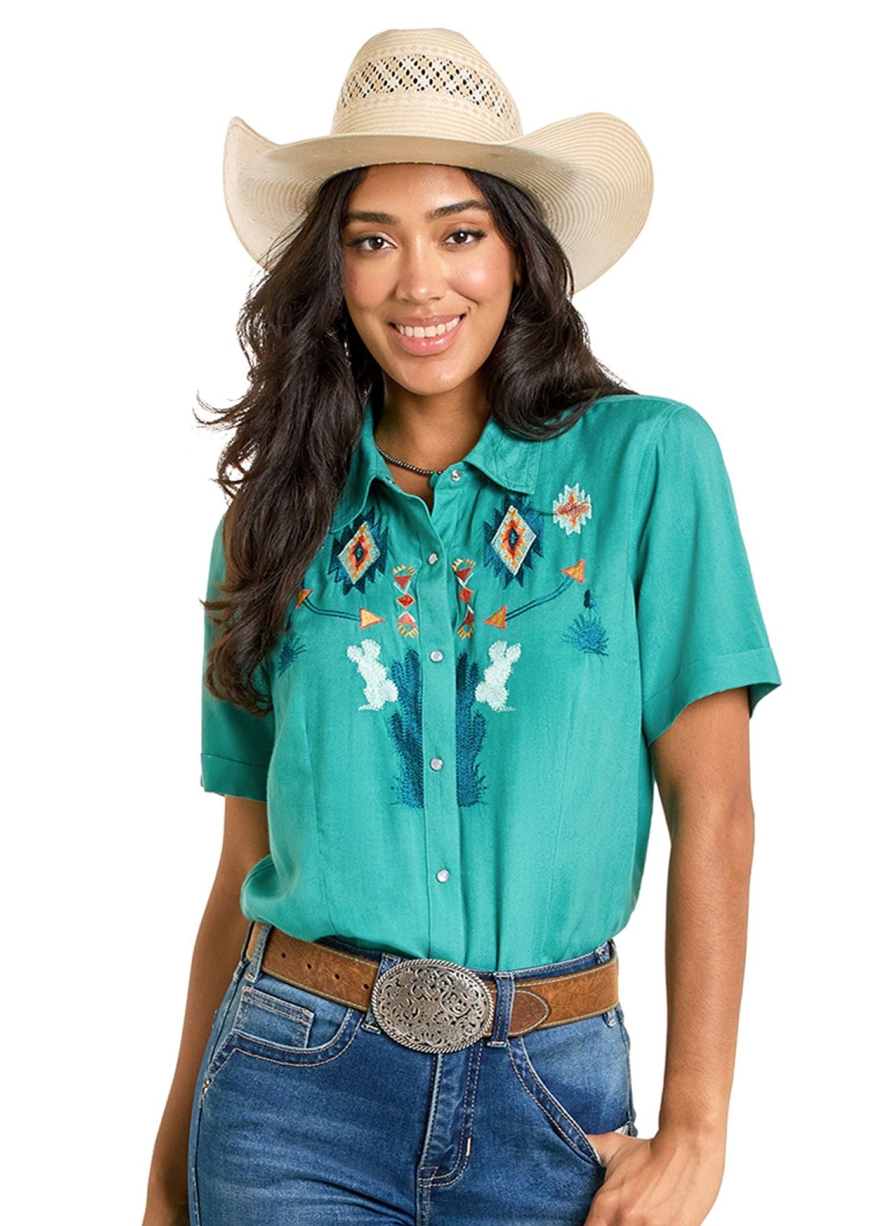 Woman wearing a teal embroidered shirt, blue jeans, and a beige cowboy hat on a white background