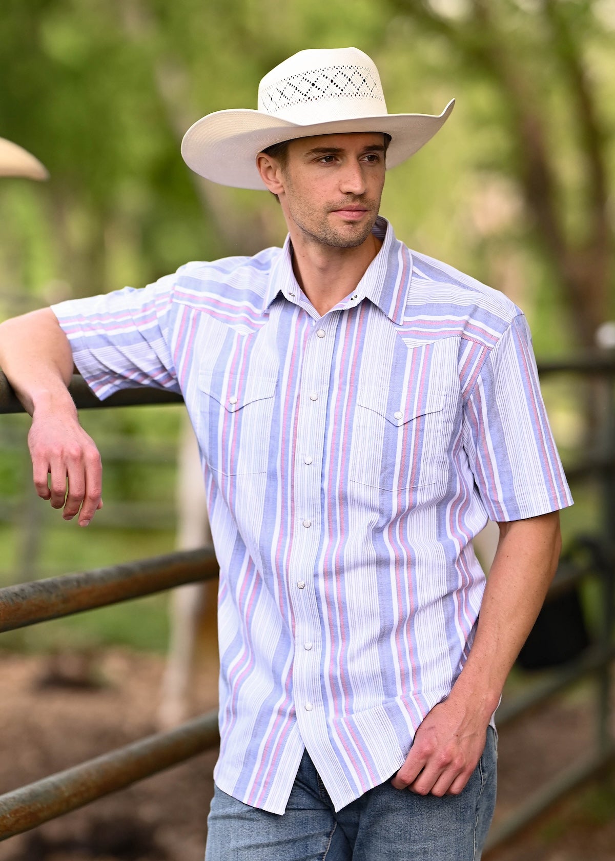 A man wearing a light blue, purple, and white striped short-sleeve western shirt, blue jeans, and a cream cowboy hat.