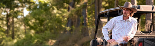 A man in a cowboy hat and striped button-down shirt leans against the back of a black off-road vehicle in a wooded outdoor setting.