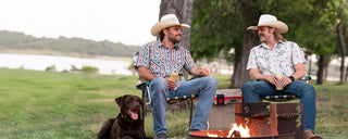 Two men in cowboy hats sitting by a fire pit with a dog near a lake.