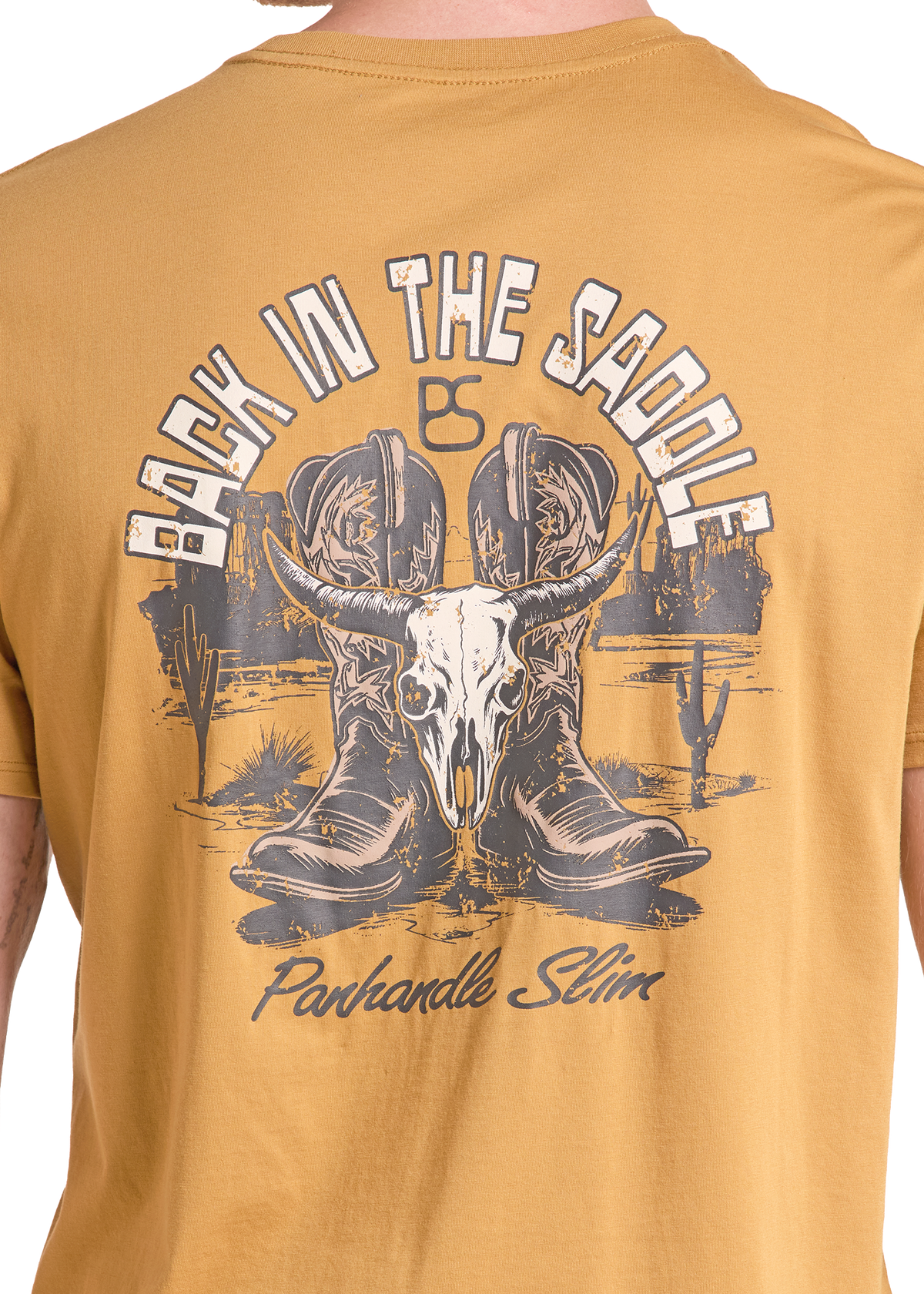 Close-up of the back of a mustard yellow t-shirt featuring a large western graphic with cowboy boots, a bull skull, cacti, and the text 'BACK IN THE SADDLE' and 'Panhandle Slim'.