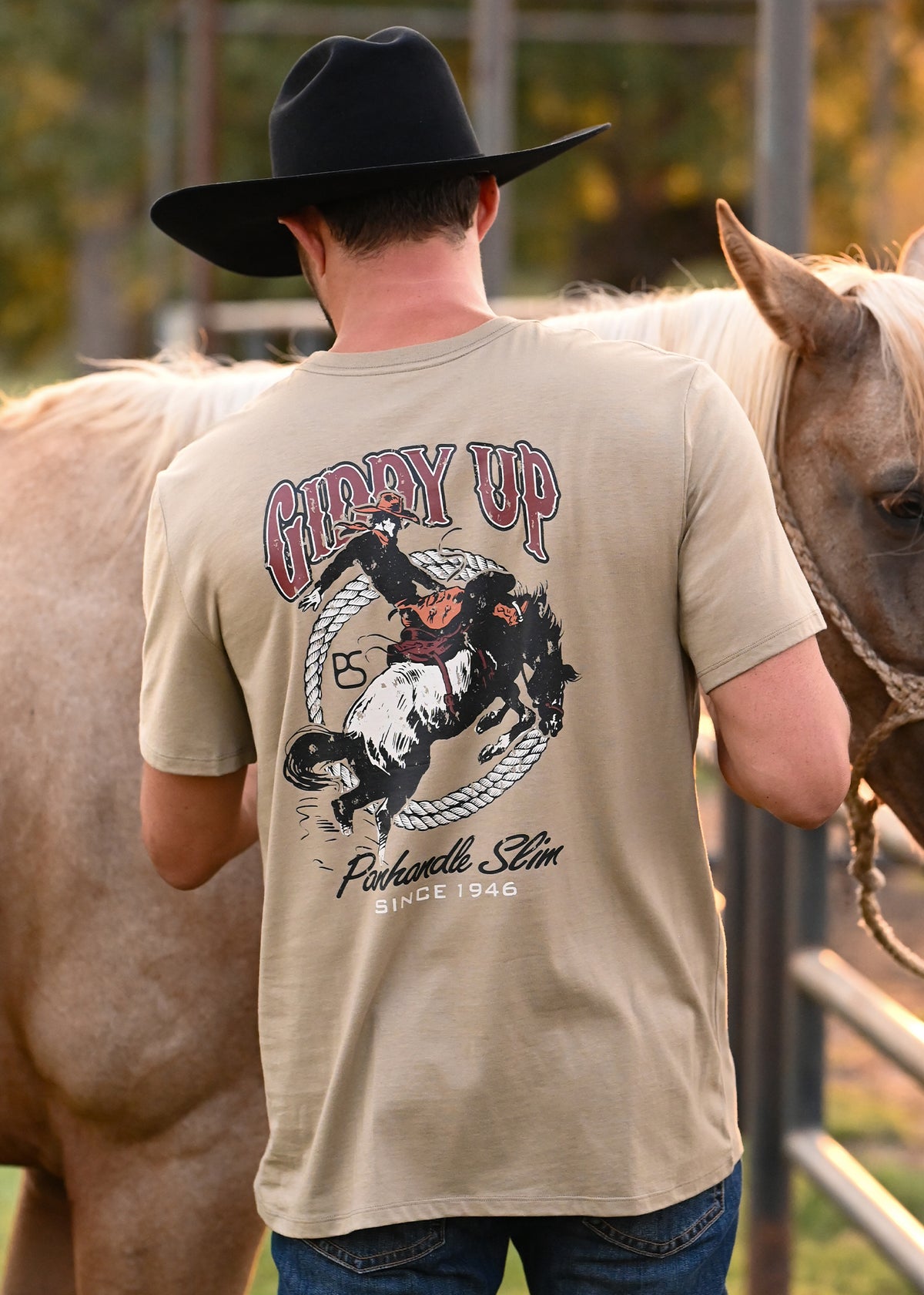 A man wearing a tan graphic t-shirt and a black cowboy hat, standing next to a horse. The back of the t-shirt features a large graphic of a cowboy riding a bucking horse.