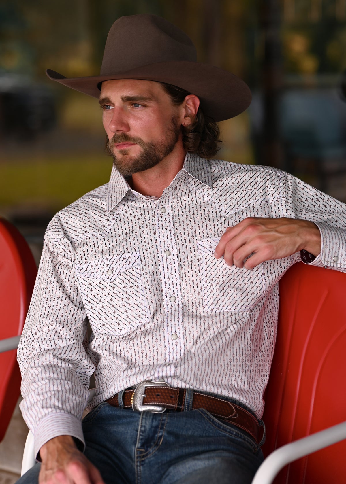 A man wearing a brown cowboy hat, a white western shirt with thin vertical brown pinstripes, and blue jeans, sitting on a red chair.