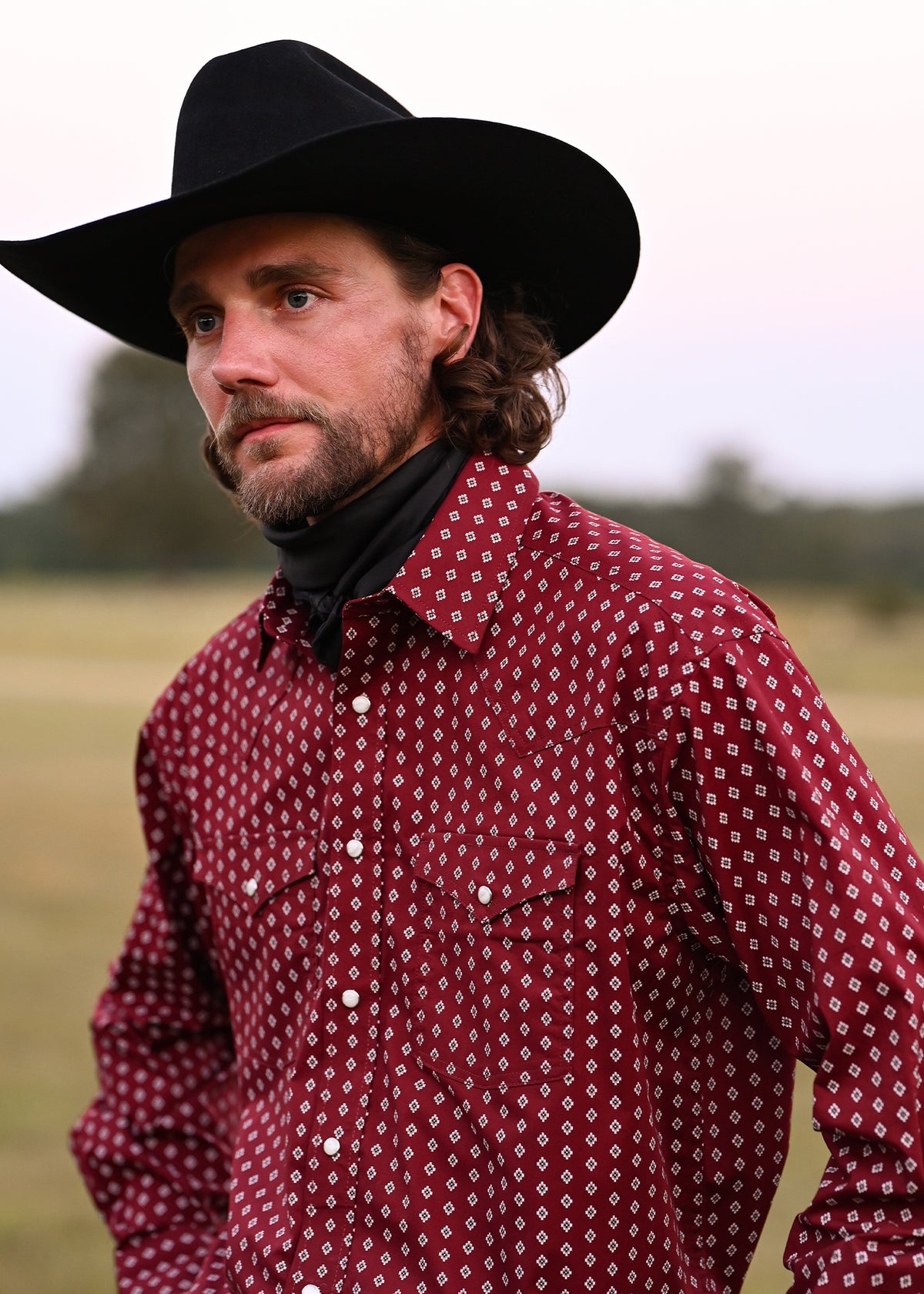 Man wearing a cowboy hat, bandana and red Western snap shirt tin a field setting. 