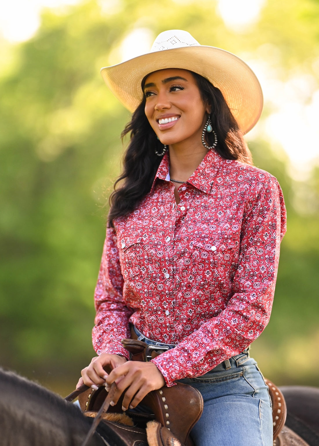 A woman wearing a red and white floral long-sleeve western shirt, blue jeans, and a beige cowboy hat. 