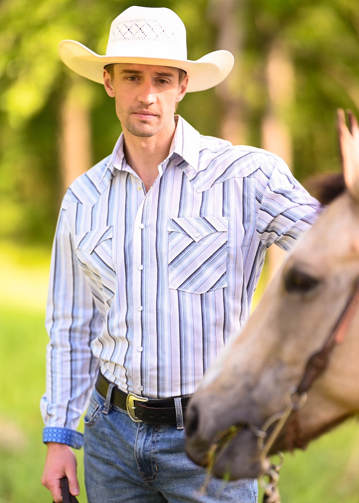 A man wearing a white cowboy hat, a blue and white striped western shirt, and jeans.