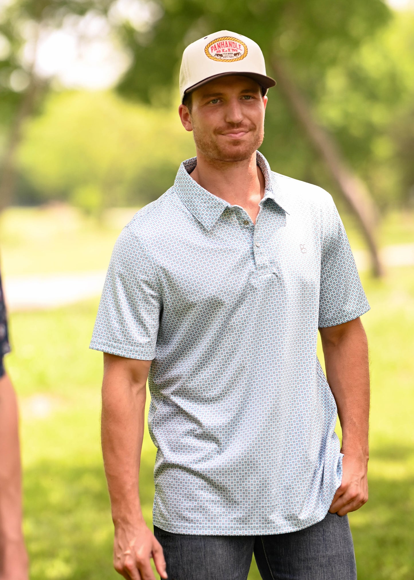 Man wearing a polo, blue jeans and a cap in a grassy setting.