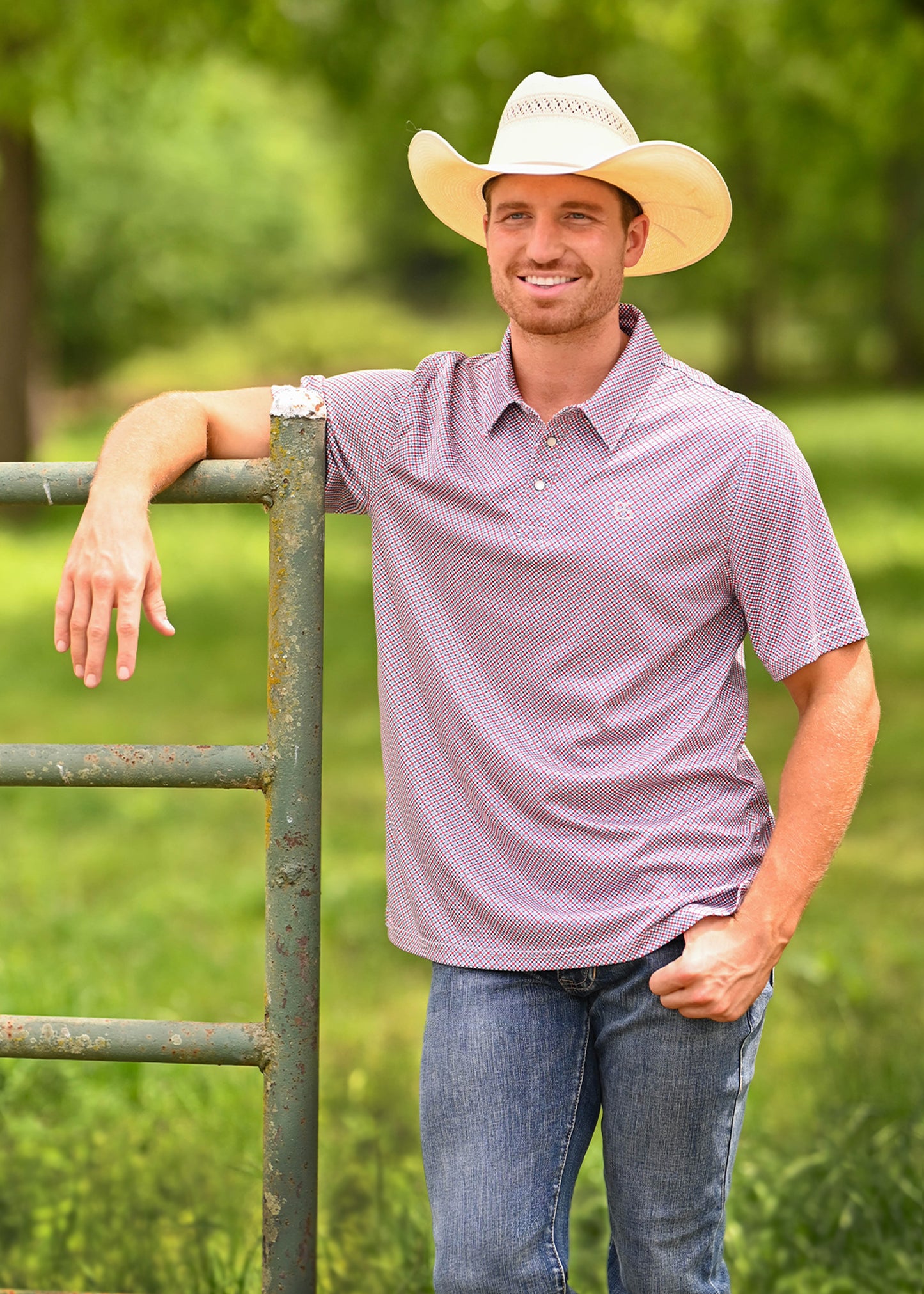 A smiling man wearing a light-colored cowboy hat, a short-sleeve button-up polo shirt with a small white geometric pattern, and blue jeans, leaning against a weathered green metal fence in a grassy outdoor setting.