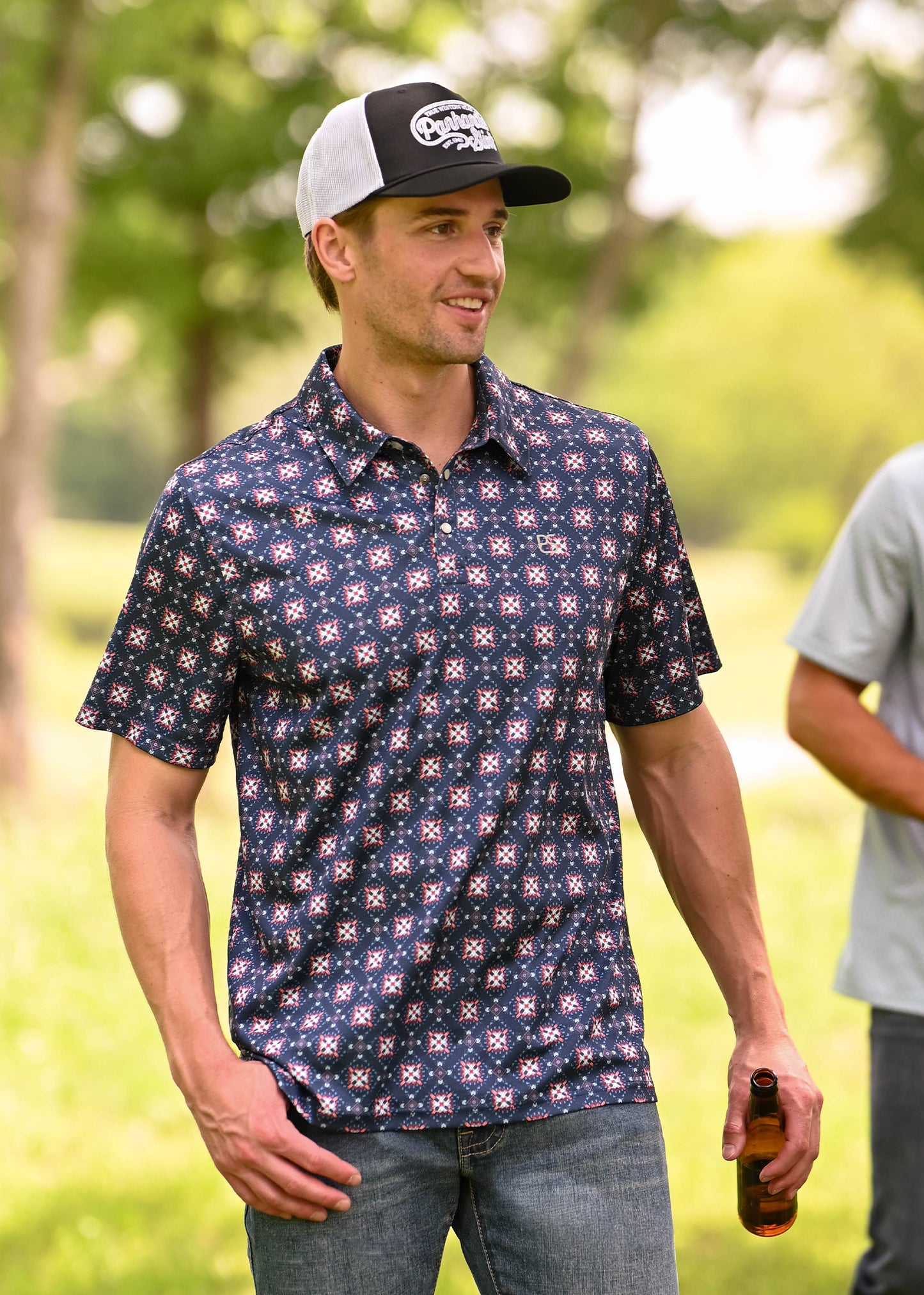 A man wearing a navy blue short-sleeve polo shirt with a white and red geometric pattern, blue jeans, and a black and white trucker hat, standing outdoors in a grassy park while holding a brown bottle.