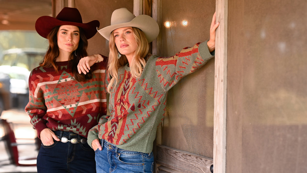 Two women wearing patterned knit sweaters and cowboy hats posing outdoors next to a rustic wooden building.