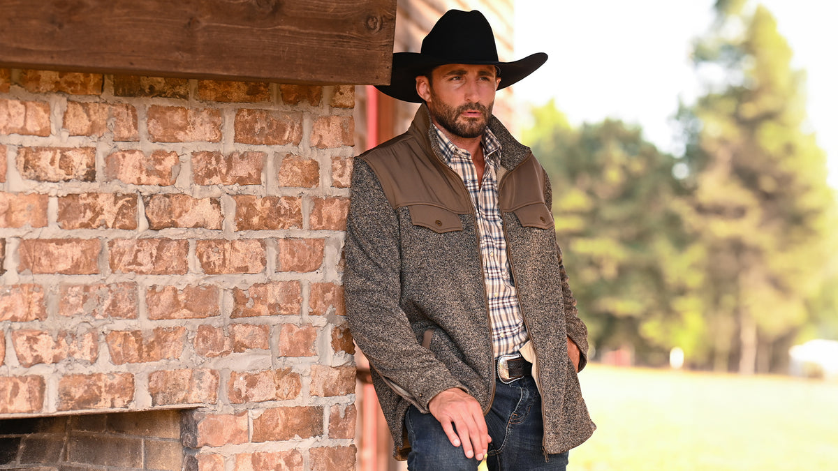 A man wearing a black cowboy hat, and a textured brown jacket over a plaid shirt, leaning against a brick wall outdoors.
